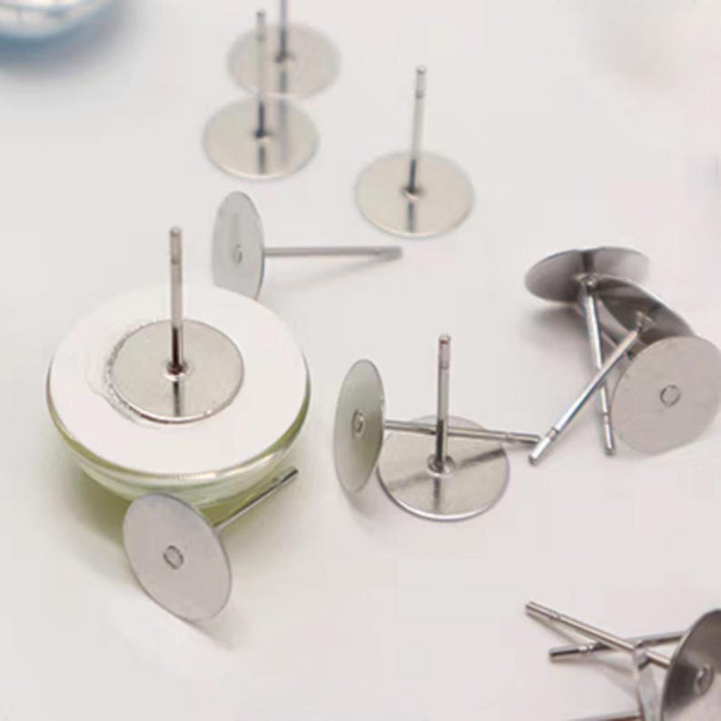 May include: Several silver earring posts with circular backings are scattered on a white surface. The posts are evenly spaced and appear to be made of metal. These components are likely for crafting or making jewellery, with a focus on earrings.