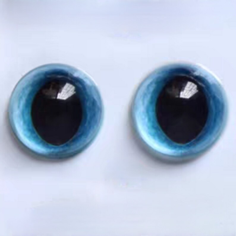 May include: Two round glass eyes with a light blue iris and a black pupil. The eyes have a reflective surface, set against a white background. These craft supplies are ideal for doll making or other artistic projects.