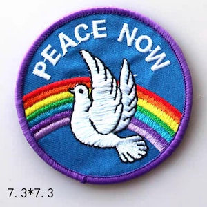 Peut inclure: Un patch rond à bordure violette avec un fond bleu. Le patch représente une colombe blanche volant au-dessus d'un arc-en-ciel avec le texte "Peace Now" en blanc.