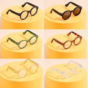 Puede incluir: Una colección de seis pares de gafas de montura redonda en varios colores. Las monturas incluyen negro, marrón, verde, carey, naranja y transparente. Cada par se muestra sobre un pedestal amarillo, mostrando los diferentes estilos y tonos.