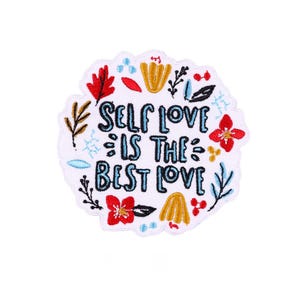 Puede incluir: Un parche bordado blanco con el texto "SELF LOVE IS THE BEST LOVE" en letras azules. El parche está decorado con diseños florales y de hojas rojas, amarillas y azules alrededor del texto.