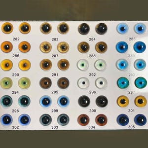 Op de afbeelding: Een verzameling poppenogen in diverse kleuren en maten, gerangschikt op een witte ondergrond. De ogen variëren van geel, bruin en groen tot blauw en zwart, elk met een glanzende afwerking. Nummers 282-305 zijn onder elk oog gedrukt.