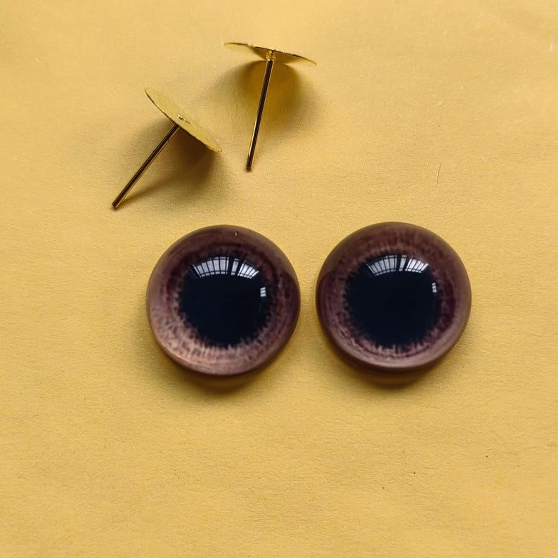 K&ouml;nnte beinhalten: Zwei Glaspuppenaugen mit einer dunkelbraunen Iris und einem hellbraunen Au&szlig;enring sowie zwei goldfarbenen Augenstiften. Die Augen haben eine reflektierende Qualit&auml;t und die Stifte haben einen flachen, kreisf&ouml;rmigen Kopf.