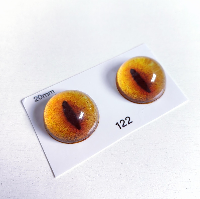 May include: Two 20mm round, glass-like eye cabochons with a yellow and brown iris and a black pupil. The eyes are on a white card with the number 122 printed on it.