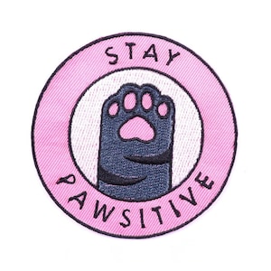 Peut inclure: Écusson brodé avec un cercle extérieur rose et un cercle intérieur blanc. Le patch présente une patte de chat bleu foncé avec un coussinet rose. Les mots "STAY PAWSITIVE" sont brodés autour de la patte.