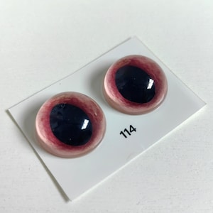 May include: Two glass doll eyes with a dark red iris and a black pupil. The eyes are set on a white card with the number 114 printed on it. The eyes have a glossy finish.