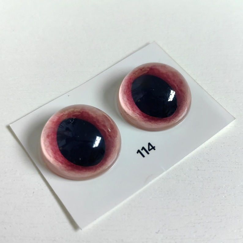 May include: Two glass doll eyes with a dark red iris and a black pupil. The eyes are set on a white card with the number 114 printed on it. The eyes have a glossy finish.