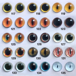 May include: A collection of glass eye cabochons in various colors and designs. The eyes feature a range of colors, including yellow, orange, brown, green, blue, and red, with black pupils. Each eye is numbered, with the numbers 117 through 135 visible.
