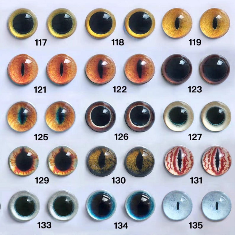 May include: A collection of glass eye cabochons in various colors and designs. The eyes feature a range of colors, including yellow, orange, brown, green, blue, and red, with black pupils. Each eye is numbered, with the numbers 117 through 135 visible.