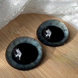 Puede incluir: Dos ojos de mu&ntilde;eca redondos de cristal con un iris azul oscuro y una pupila negra. Los ojos tienen una superficie reflectante y brillante y est&aacute;n colocados sobre un fondo de madera. El borde exterior de los ojos es azul claro.