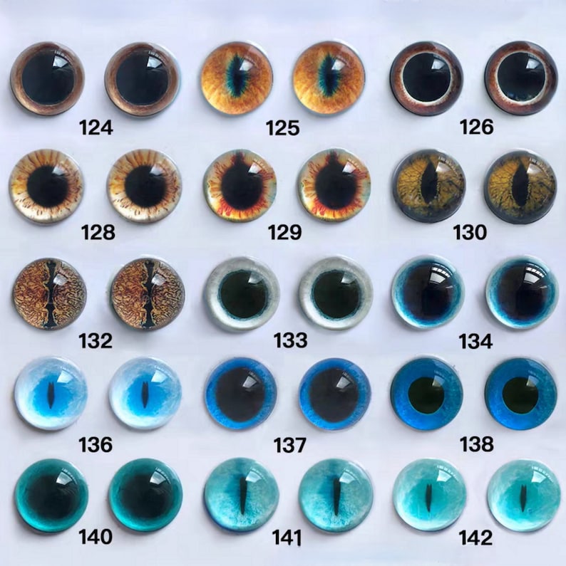 May include: A collection of glass eye cabochons in various colors and designs. The eyes feature a range of colors, including brown, orange, yellow, green, blue, and teal. Each eye has a black pupil and is numbered for identification.