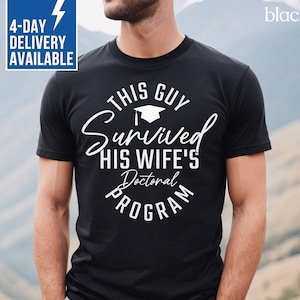 May include: Black t-shirt with a white graphic that says "This Guy Survived His Wife's Doctoral Program" with a graduation cap icon.