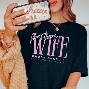 May include: Black t-shirt with the text "Pastor's Wife" in pink lettering. Below the text is "Cross Church Fayetteville, AR".