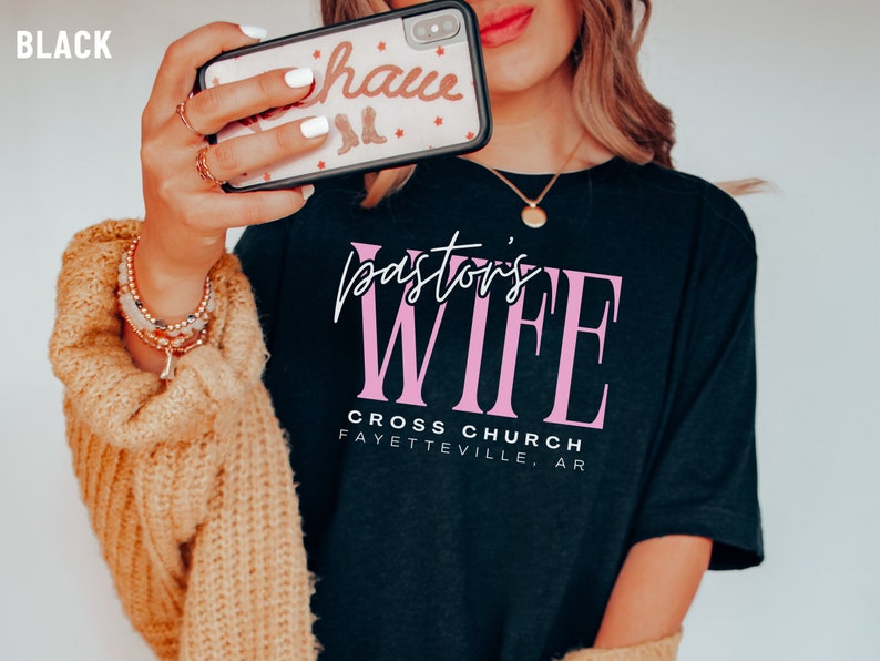 May include: Black t-shirt with the text "Pastor's Wife" in pink lettering. Below the text is "Cross Church Fayetteville, AR".