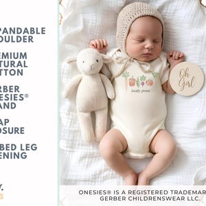 May include: A newborn baby wearing a cream-colored onesie with a vegetable graphic and the words "locally grown." The baby is lying in a woven basket with a stuffed lamb and a wooden disc that says "Oh Girl."