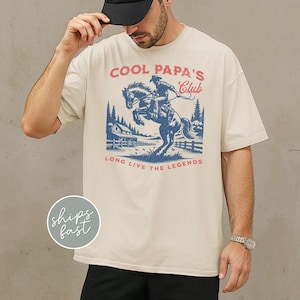 May include: Ivory-colored t-shirt with a graphic of a cowboy riding a horse, with the text "COOL PAPA'S Club" and "LONG LIVE THE LEGENDS" in red and blue. The design includes a ranch scene. A black cap is worn.
