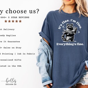 Puede incluir: Camiseta azul denim con la frase "It's fine. I'm fine. Everything's fine." y un dise&ntilde;o de mapache sosteniendo un cuchillo y un gato. La camiseta es de corte holgado.
