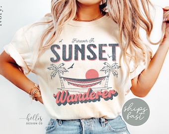 May include: Ivory-colored t-shirt with a graphic design featuring the words "Forever A Sunset Wanderer." The design includes a red sun, palm trees, and a hammock. The shirt has a relaxed fit.