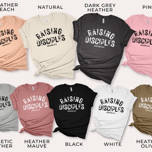 May include: A collection of t-shirts in various colors featuring the text "Raising Disciples" with the scripture reference "Proverbs 22:6". The shirts are available in Heather Peach, Natural, Dark Grey Heather, Pink, Athletic Heather, Heather Mauve, Black, White, and Heather Olive.