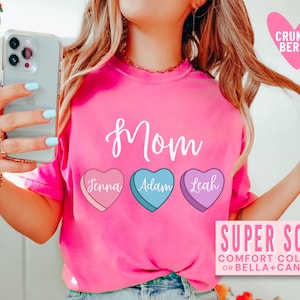 May include: Pink t-shirt with the word "Mom" in white script and three candy hearts with the names "Jenna", "Adam", and "Leah" printed on them. The text "SUPER SOFT COMFORT COLORS OR BELLA+CANVAS" is printed at the bottom of the shirt.
