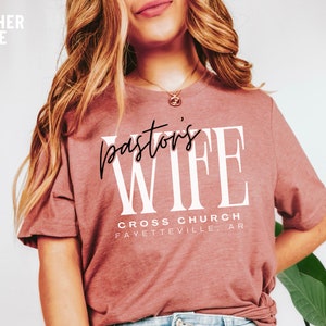 May include: A heather mauve t-shirt with the text "pastor's wife" in white lettering. The text "CROSS CHURCH FAYETTEVILLE, AR" is printed below in black lettering.