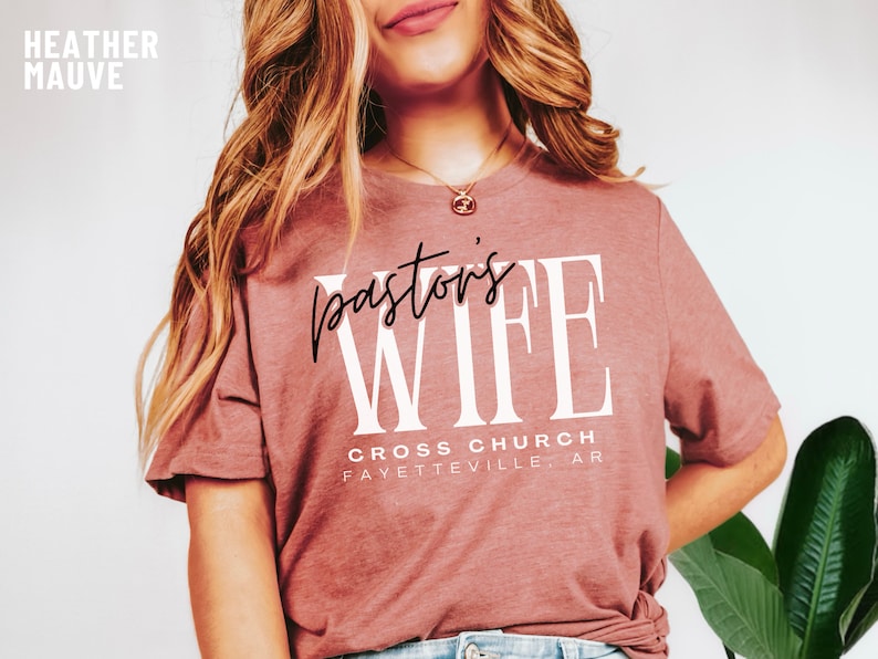 May include: A heather mauve t-shirt with the text "pastor's wife" in white lettering. The text "CROSS CHURCH FAYETTEVILLE, AR" is printed below in black lettering.