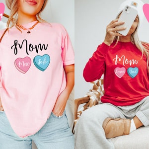 May include: Two pink and red t-shirts with the word "Mom" and two candy hearts with names on them. The pink shirt has the names "Mac" and "Josh" on the hearts. The red shirt has the names "Emma" and "Fred" on the hearts.