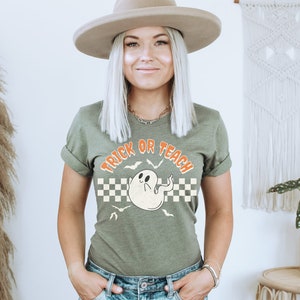 Op de afbeelding: Een groen T-shirt met een witte geest en de tekst "Trick or Teach" in oranje. De geest heeft een geruit patroon erachter en vleermuizen vliegen eromheen.