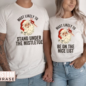May include: Two white t-shirts featuring a vintage illustration of Santa Claus. The text on the shirts reads "Most Likely To" with a phrase below. One shirt says "Stand Under The Mistletoe" and the other says "Be On The Nice List".