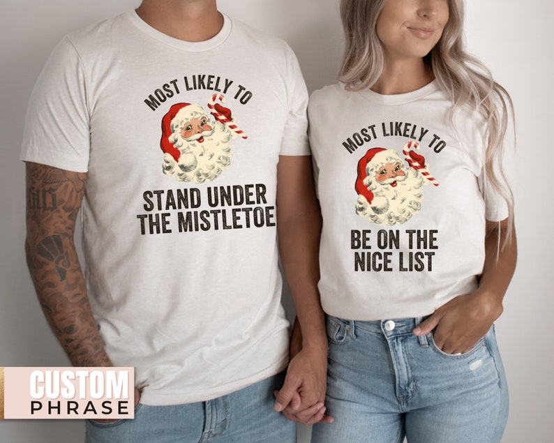 May include: Two white t-shirts featuring a vintage illustration of Santa Claus. The text on the shirts reads "Most Likely To" with a phrase below. One shirt says "Stand Under The Mistletoe" and the other says "Be On The Nice List".