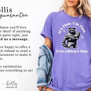 Puede incluir: Camiseta violeta con la frase "It's fine. I'm fine. Everything's fine." y un gr&aacute;fico de un mapache sosteniendo un gato. Tambi&eacute;n incluye el texto "hollis guarantee".