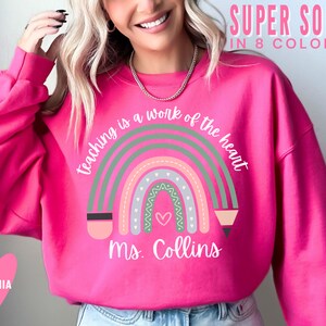 May include: Pink sweatshirt with a rainbow design and the text "teaching is a work of the heart" and "Ms. Collins".