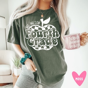 K&ouml;nnte beinhalten: Ein dunkelgr&uuml;nes T-Shirt mit einem wei&szlig;en Apfel-Motiv, auf dem in einer Retro-Schrift "Ms. Wilson Fourth Grade" steht. Der Apfel hat ein kariertes Muster.