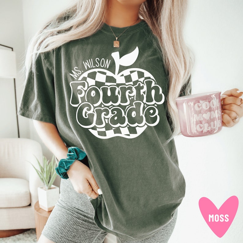 K&ouml;nnte beinhalten: Ein dunkelgr&uuml;nes T-Shirt mit einem wei&szlig;en Apfel-Motiv, auf dem in einer Retro-Schrift "Ms. Wilson Fourth Grade" steht. Der Apfel hat ein kariertes Muster.