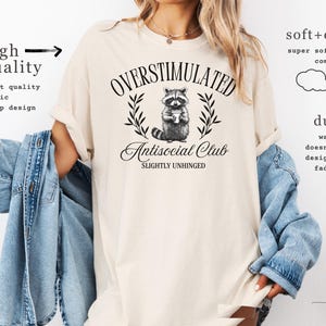 Puede incluir: Camiseta color marfil con el texto "OVERSTIMULATED Antisocial Club SLIGHTLY UNHINGED" y un gr&aacute;fico de mapache. La camiseta est&aacute; hecha de una tela suave y c&oacute;moda y tiene un dise&ntilde;o n&iacute;tido.