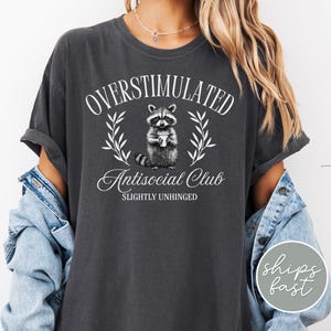 Puede incluir: Camiseta gris oscuro con las palabras blancas "OVERSTIMULATED Antisocial Club SLIGHTLY UNHINGED". Una ilustraci&oacute;n de mapache est&aacute; centrada en la camiseta. La camiseta tiene las mangas enrolladas y se usa con una chaqueta vaquera.