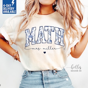 May include: Ivory-colored t-shirt with the word "MATH" in blue floral letters. Below the word is "mrs. miller" in cursive, with a small heart. The shirt is a crew neck and has short sleeves.