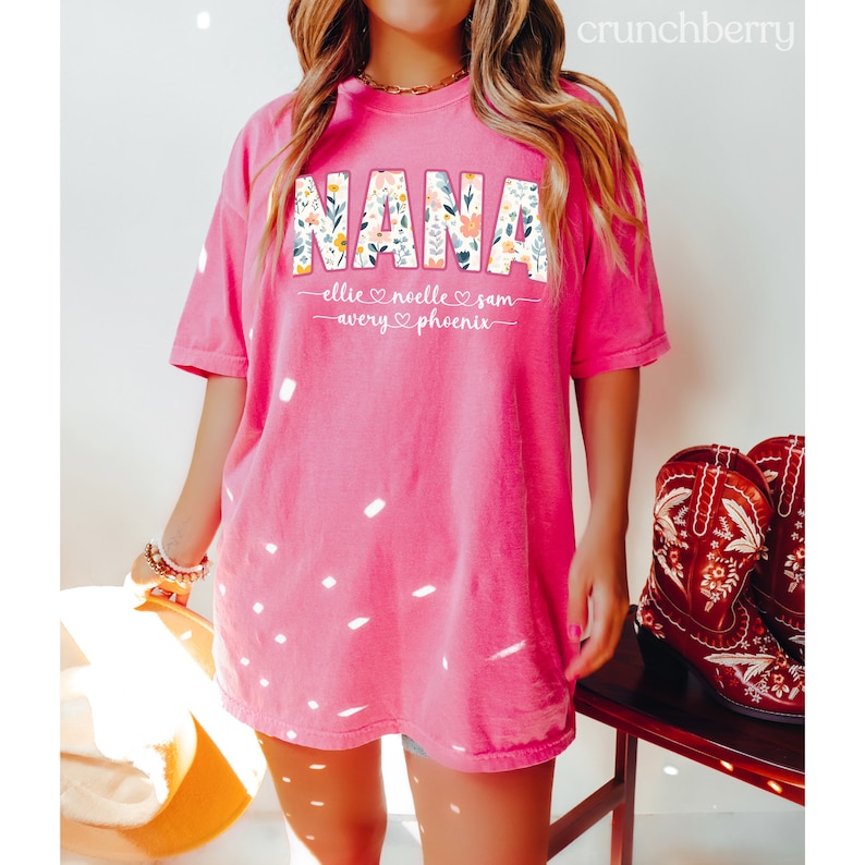 K&ouml;nnte beinhalten: Rosafarbenes Oversize-T-Shirt mit dem Wort "NANA" in Blumenbuchstaben. Das Shirt tr&auml;gt die Namen "Ellie, Noelle, Sam, Avery, Phoenix" unter dem Wort "NANA" mit Herzen zwischen den Namen.