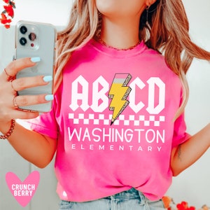 May include: Pink t-shirt with a white graphic that says "ABCD" with a yellow lightning bolt and a checkered pattern. The text "WASHINGTON ELEMENTARY" is below the graphic.