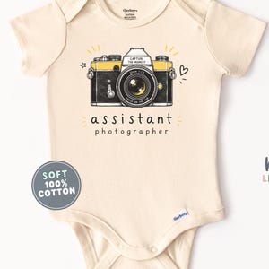 May include: A cream-colored short-sleeve bodysuit featuring a graphic of a black and yellow camera with the text "CAPTURE THE MOMENT" and "FOCUS ON THE FUN" on the lens, with the words "assistant photographer" printed below the camera.