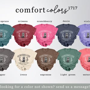 Puede incluir: Una colecci&oacute;n de camisetas Comfort Colors en varios tonos, incluyendo azul abeto, carmes&iacute; y violeta. Cada camiseta muestra el texto "OVERSTIMULATED Antisocial Club" con un gr&aacute;fico de mapache. La imagen tambi&eacute;n incluye el texto "comfort colors 1717".