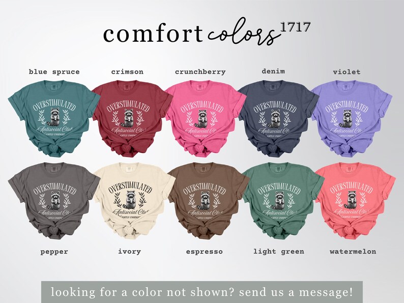 Puede incluir: Una colecci&oacute;n de camisetas Comfort Colors en varios tonos, incluyendo azul abeto, carmes&iacute; y violeta. Cada camiseta muestra el texto "OVERSTIMULATED Antisocial Club" con un gr&aacute;fico de mapache. La imagen tambi&eacute;n incluye el texto "comfort colors 1717".