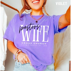 May include: A purple t-shirt with the text "Pastor's Wife" in white letters and "Cross Church Fayetteville, AR" in black letters.