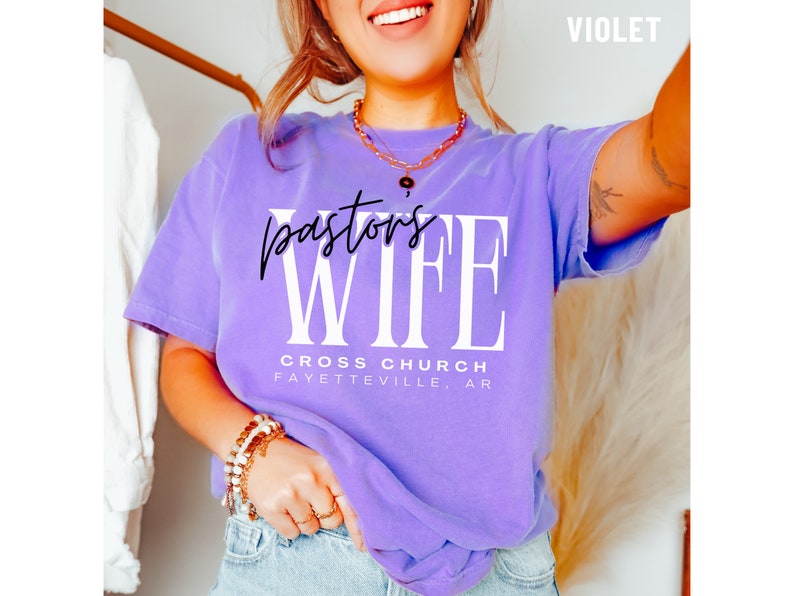May include: A purple t-shirt with the text "Pastor's Wife" in white letters and "Cross Church Fayetteville, AR" in black letters.
