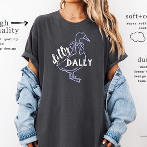 May include: Dark grey t-shirt featuring a white and purple duck graphic with the words "dilly dally". The shirt has rolled sleeves and is made of a soft, comfortable fabric.