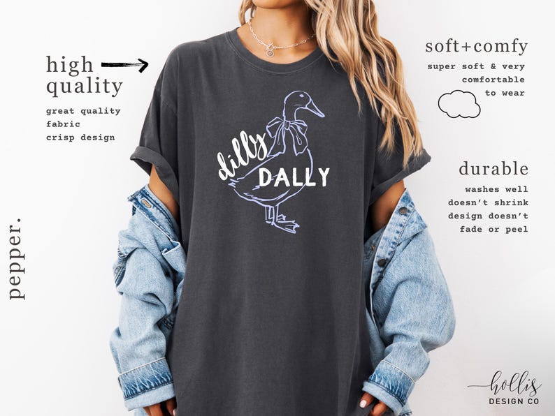 May include: Dark grey t-shirt featuring a white and purple duck graphic with the words "dilly dally". The shirt has rolled sleeves and is made of a soft, comfortable fabric.
