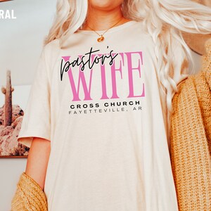 May include: A white t-shirt with the text "Pastor's Wife" in black and pink lettering. The text "Cross Church Fayetteville, AR" is printed below in black.