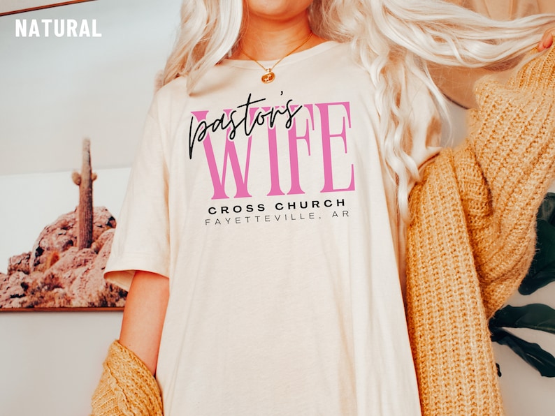 May include: A white t-shirt with the text "Pastor's Wife" in black and pink lettering. The text "Cross Church Fayetteville, AR" is printed below in black.