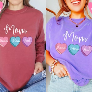 May include: Two long-sleeved shirts with the word "Mom" and three candy hearts with names "Jenna", "Adam", and "Leah". The shirt on the left is crimson and the shirt on the right is violet.