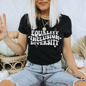 以下が含まれることがあります： レトロなフォントで「Equality Inclusion Diversity」と書かれた黒いTシャツを着た女性。彼女は、かぼちゃや秋の装飾品が置かれた白い表面に座っています。